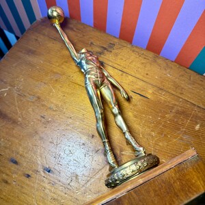 Puede incluir: Trofeo de baloncesto dorado que presenta una figura estilizada con ropa deportiva, extendiendo la mano para sostener un bal&oacute;n de baloncesto. El trofeo est&aacute; montado sobre una base decorativa y es de metal brillante.