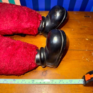 May include: Close-up of a vintage doll's legs. The doll's legs are covered in red, fuzzy fabric, and the feet are black, glossy, and rounded. The legs are positioned on a wooden surface.