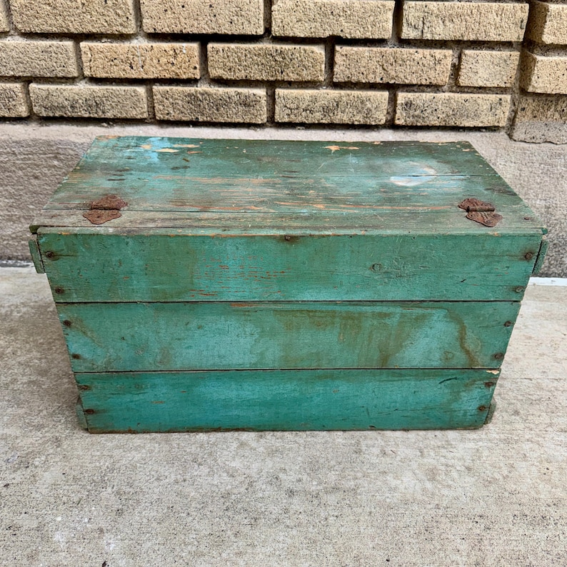May include: A weathered, rectangular wooden box painted in a distressed teal color. The box has a hinged lid with two rusty metal hinges. The wood grain is visible, and the box appears to be vintage or antique.