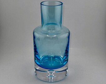 Könnte beinhalten: Eine transparente, türkisfarbene Glasvase mit zylindrischem Hals und rundem Körper. Die Vase steht auf einem transparenten, kreisförmigen Sockel. Das Glas hat eine glatte, glänzende Oberfläche. Die Vase ist leer. Höhe ca. 15 cm.