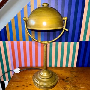 Puede incluir: Una lámpara de escritorio de latón de estilo vintage con una pantalla en forma de cúpula. La lámpara tiene un brazo curvo y una base redonda. La pantalla y la base tienen un acabado dorado antiguo y cálido. La lámpara está sobre una superficie de madera con un fondo a rayas de colores.
