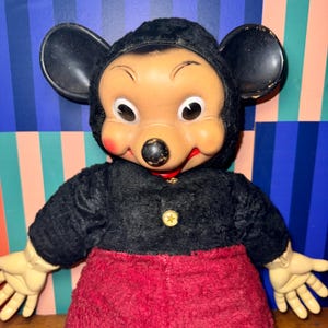 May include: Vintage Mickey Mouse doll with a black plush coat, red pants, and large black ears. The doll has a tan face with rosy cheeks, a black nose, and a red mouth. A gold star-shaped button adorns the coat.