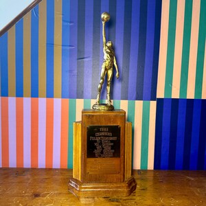 Puede incluir: Un trofeo dorado vintage que representa a un jugador de baloncesto sosteniendo un bal&oacute;n en alto. El trofeo est&aacute; montado sobre una base de madera con una placa que dice "1949 Champions". El fondo tiene rayas verticales coloridas.
