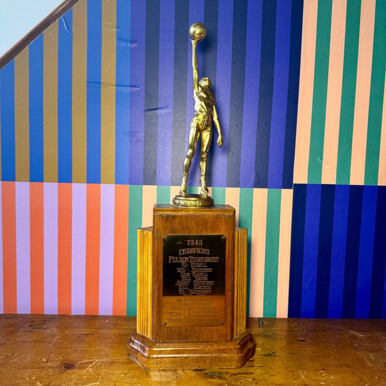 Puede incluir: Un trofeo dorado vintage que representa a un jugador de baloncesto sosteniendo un bal&oacute;n en alto. El trofeo est&aacute; montado sobre una base de madera con una placa que dice "1949 Champions". El fondo tiene rayas verticales coloridas.