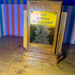 Puede incluir: Un trofeo de madera vintage con una placa dorada. La placa dice "1949 CHAMPIONS FOLSOM TOURNAMENT" con una lista de nombres. El trofeo tiene un acabado de madera oscura y un dise&ntilde;o decorativo.