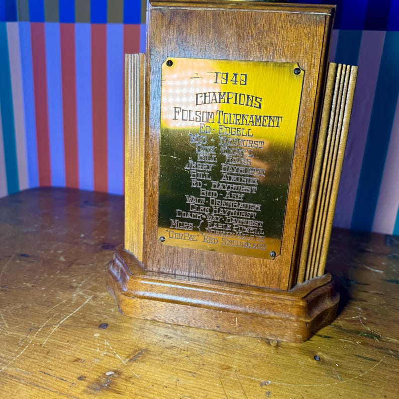 Puede incluir: Un trofeo de madera vintage con una placa dorada. La placa dice "1949 CHAMPIONS FOLSOM TOURNAMENT" con una lista de nombres. El trofeo tiene un acabado de madera oscura y un dise&ntilde;o decorativo.