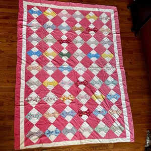 Op de afbeelding: Een handgemaakte quilt met een roze achtergrond en een witte rand. De quilt heeft een ruitpatroon met afwisselend roze en witte vierkanten. Elk wit vierkant bevat een kleurrijk vlinderdasontwerp in verschillende kleuren.