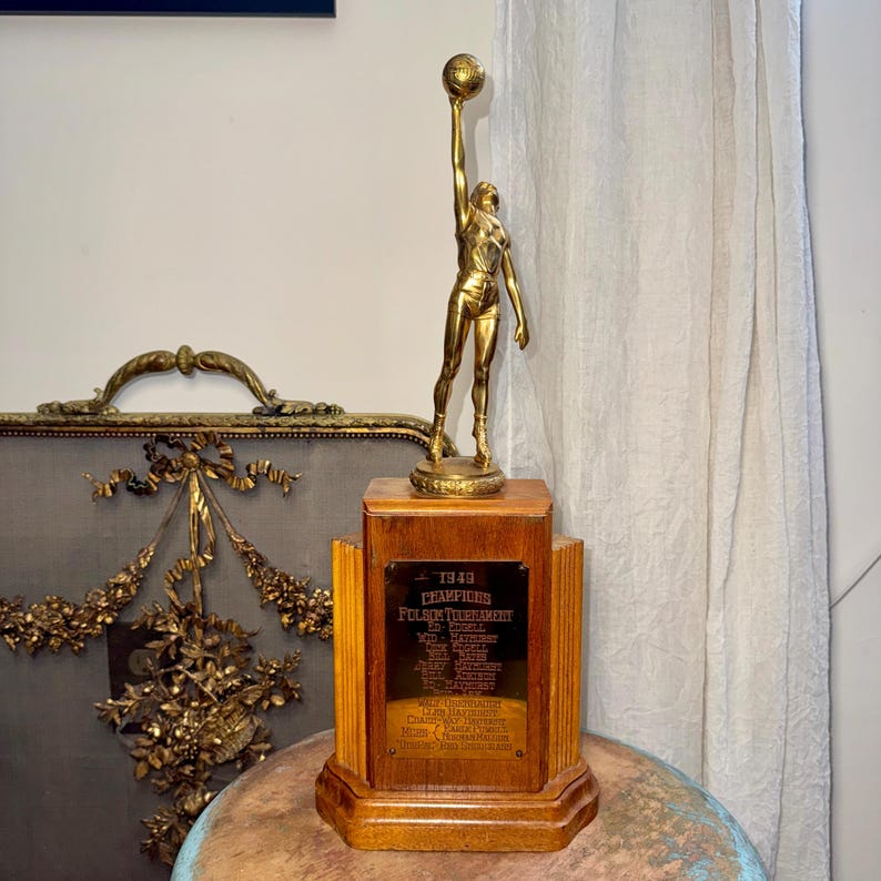 Puede incluir: Un trofeo de baloncesto vintage con una figura dorada sosteniendo un bal&oacute;n por encima de su cabeza. La figura est&aacute; montada sobre una base de madera con una placa de lat&oacute;n. La placa dice "1949 Champions".