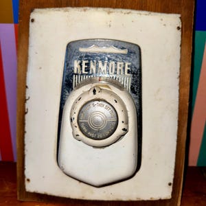 Op de afbeelding: Vintage Kenmore apparaat bedieningspaneel. Het paneel is wit met een chroomkleurig bovendeel met de tekst "KENMORE". Een ronde draaiknop met instructies is gecentreerd onder de merknaam. Het paneel is op een houten achterkant gemonteerd.