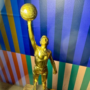 Puede incluir: Una estatua dorada de un jugador de baloncesto sosteniendo un bal&oacute;n por encima de la cabeza. La figura lleva una camiseta sin mangas y pantalones cortos. El fondo tiene rayas verticales azules, naranjas y verdes.