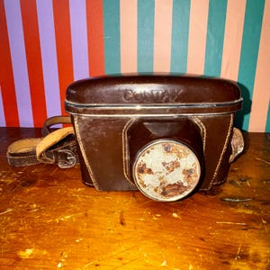May include: A vintage brown leather camera case with the brand name "CONTAX" embossed on the top. The case has a worn appearance, with a circular lens opening and a leather strap. The camera case is sitting on a wooden surface.