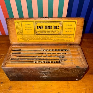 May include: An open wooden box containing a set of spur auger bits. The box has a yellow label with black text that reads "RUSSELL JENNINGS SPUR AUGER BITS." The bits are arranged in individual slots within the box.