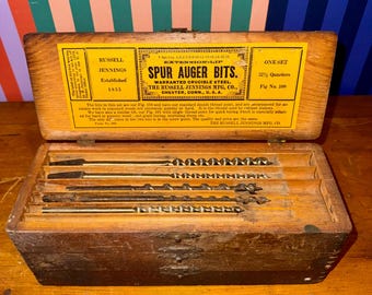 May include: An open wooden box containing a set of spur auger bits. The box has a yellow label with black text that reads "RUSSELL JENNINGS SPUR AUGER BITS." The bits are arranged in individual slots within the box.