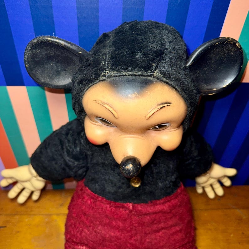 May include: Vintage Mickey Mouse doll with a black plush hood and body, red pants, and tan hands. The doll has a painted face with a black nose and a gold-colored charm on the chest. The doll's expression is slightly stern.