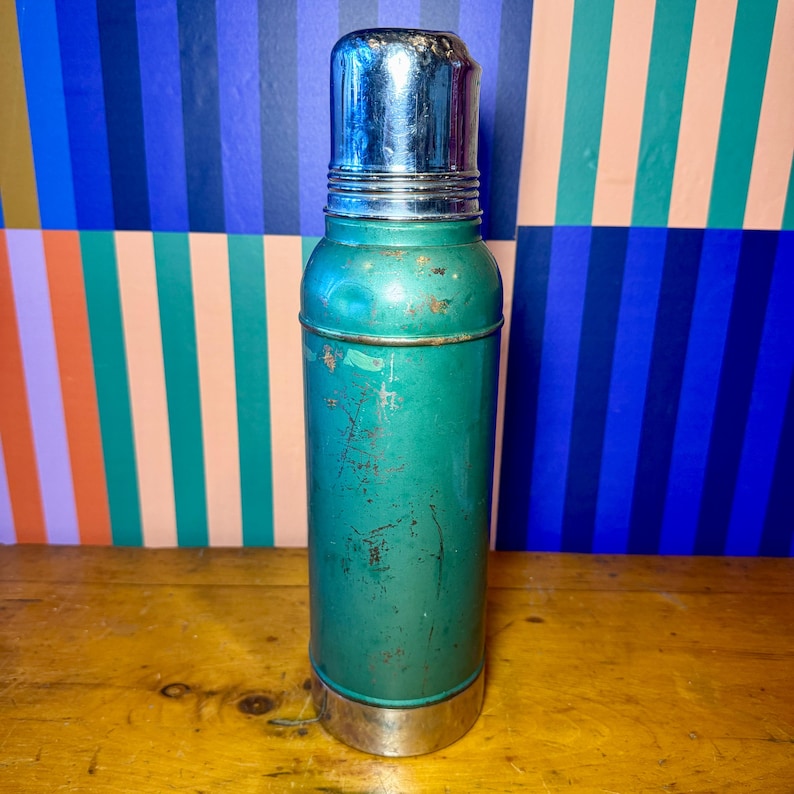 Pode incluir: Termo de metal verde vintage com tampa e base prateadas brilhantes. O termo tem formato cil&iacute;ndrico e apresenta sinais de desgaste. O fundo apresenta um padr&atilde;o de listras coloridas.