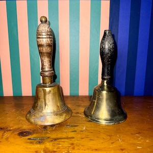 May include: Two antique brass bells with wooden handles. One bell has a light brown handle and the other has a black handle. The bells are on a wooden surface with a striped background of pink, green, and blue.