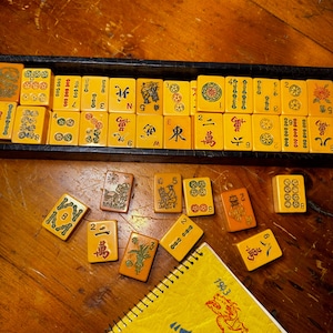 Op de afbeelding: Een vintage Mahjong-set met gele tegels, sommige gerangschikt in een zwarte lade, andere verspreid op een houten oppervlak. De tegels bevatten Chinese karakters en symbolen. Een geel notitieblok met het jaar 1943 is ook zichtbaar.