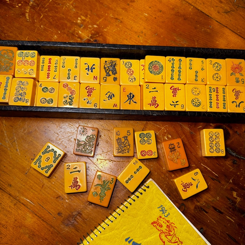 Op de afbeelding: Een vintage Mahjong-set met gele tegels, sommige gerangschikt in een zwarte lade, andere verspreid op een houten oppervlak. De tegels bevatten Chinese karakters en symbolen. Een geel notitieblok met het jaar 1943 is ook zichtbaar.