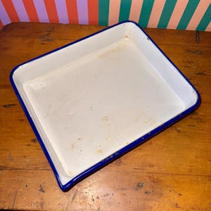 May include: A rectangular white enamel baking dish with a blue rim. The dish has a slightly worn appearance, with some discoloration on the white surface. The blue rim provides a contrasting border to the white interior.