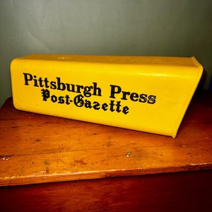 Peut inclure: Un conteneur rectangulaire jaune avec le texte "Pittsburgh Press Post-Gazette" en lettres noires. Le conteneur est posé sur une surface en bois de couleur brun rougeâtre.
