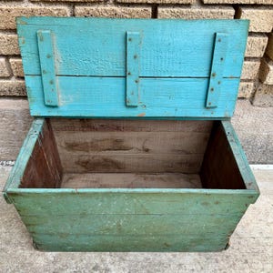 May include: An open, weathered wooden storage chest with a hinged lid. The exterior is painted in shades of turquoise and green, with visible wood grain. The interior is unpainted, showing the natural wood. The chest is rectangular.