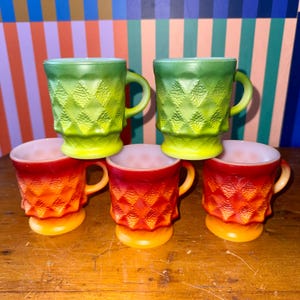 May include: Five vintage glass mugs in shades of green and red-orange. The mugs have a diamond pattern and a handle. The green mugs are on top, and the red-orange mugs are below. The mugs are on a wooden surface.