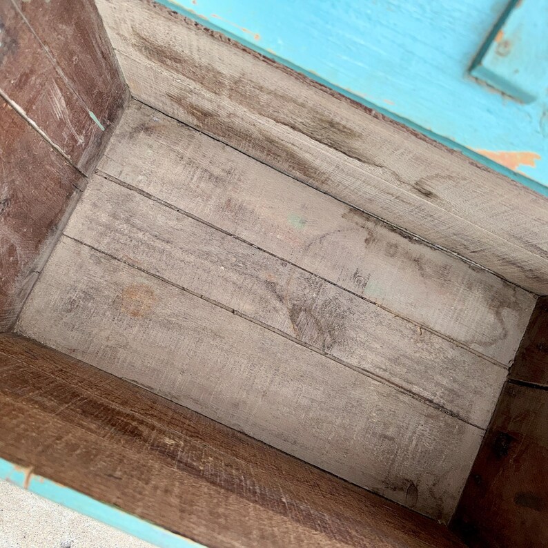 May include: An open wooden box with a distressed, weathered appearance. The interior features light gray planks, while the sides and top edges are painted in a vibrant turquoise. The wood grain is visible throughout, adding to the rustic charm.