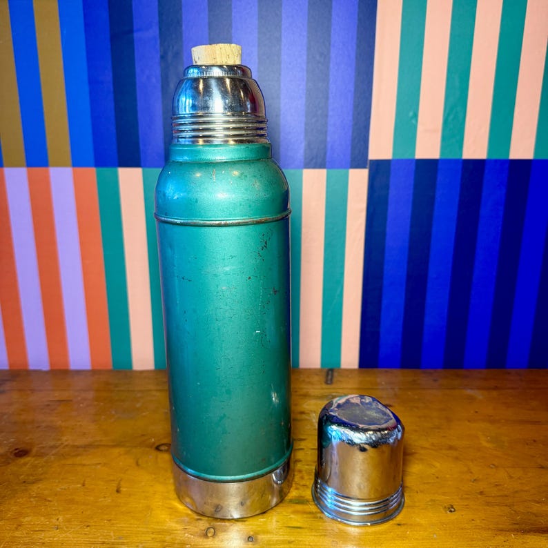 Pode incluir: Termo vintage verde-azulado com tampa e base prateadas e rolha de corti&ccedil;a. O termo &eacute; cil&iacute;ndrico e tem um aspeto ligeiramente desgastado. A tampa est&aacute; separada e fica ao lado do termo. O fundo &eacute; um padr&atilde;o de riscas coloridas.