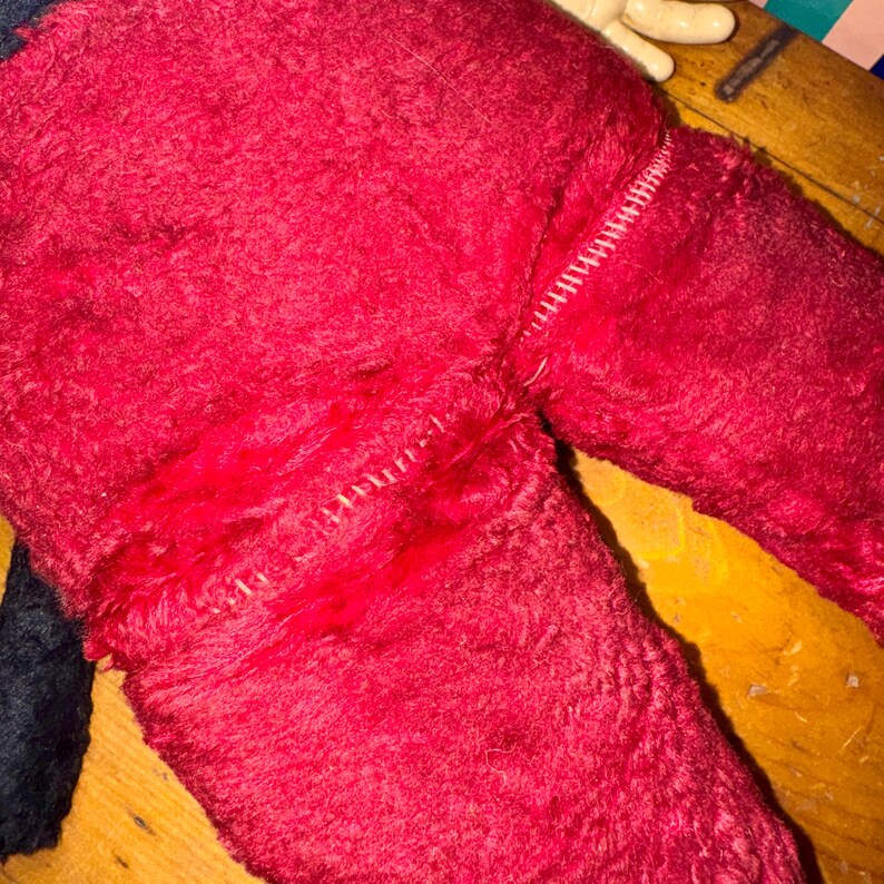 May include: Close-up of a red plush toy with visible stitching. The toy's fur is a deep red, with a lighter red patch and white stitching. A portion of a black fabric is visible on the left side.