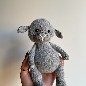 May include: A grey crocheted stuffed lamb toy with black eyes and a white nose. The lamb is sitting in someone's hand.