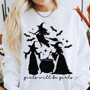 Może przedstawiać: Biała bluza z czarną grafiką przedstawiającą trzy wiedźmy, nietoperze i gwiazdy. Pod wiedźmami wydrukowano napis "girls will be girls". Motyw Halloween.