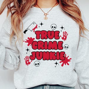 May include: A light gray sweatshirt with the text "TRUE CRIME JUNKIE" in red, with a black cleaver, red handprints, skulls, and star accents. The sweatshirt is a casual style.