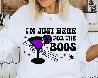 May include: White sweatshirt with the text "I'm just here for the boos" in black lettering. A purple cocktail with a lime wedge and a skeleton hand holding the glass is pictured below the text. A black spider is in the upper right corner of the design.
