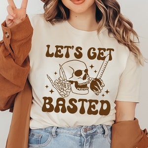 Puede incluir: Una camiseta blanca con un gráfico marrón de un esqueleto sosteniendo una jeringa y una regla. El texto "Let's Get Basted" está impreso en letras marrones.
