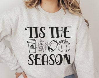 May include: A gray sweatshirt with the text 'Tis the season' in black. The text is surrounded by four black icons: a coffee cup with a pumpkin inside, a leaf, a football, and a pumpkin.