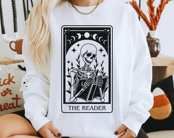 Può includere: Felpa bianca con un'illustrazione in bianco e nero di uno scheletro che legge un libro. L'illustrazione è in stile carta dei tarocchi e dice "The Reader" in basso.