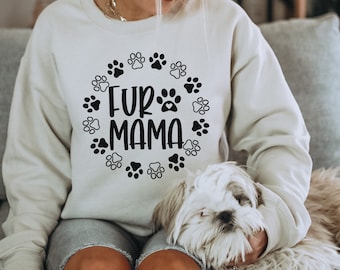 以下が含まれることがあります： 黒い肉球のデザインと「Fur Mama」という文字が円形に書かれた白いスウェットシャツ。白い小型犬が、スウェットシャツを着た人の膝の上に座っている。