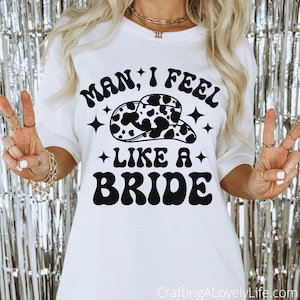 以下が含まれることがあります： 白いTシャツに、黒い文字で「Man, I feel like a bride」と書いてあり、牛柄のテンガロンハットのグラフィックが描かれている。