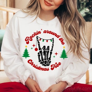 Puede incluir: Sudadera blanca con una mano de esqueleto negra haciendo un signo de rock and roll con luces de Navidad enhebradas alrededor de los dedos. El texto "Rockin' around the Christmas Tree" está escrito en rojo y blanco.