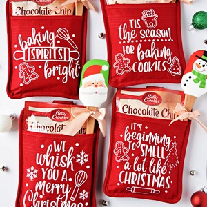 Può includere: Quattro guanti da forno rossi con diversi detti a tema natalizio e una scatola di mix per biscotti al cioccolato Betty Crocker e una spatola di legno con un design natalizio. I detti sono "baking spirits bright", "Tis the season for baking cookies", "It's beginning to smell a lot like Christmas", e "We whisk you a merry Christmas".