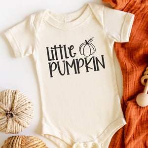 May include: A white baby onesie with the text "Little Pumpkin" and a black outline of a pumpkin. The onesie is lying on a white surface with a brown blanket and two woven pumpkins.