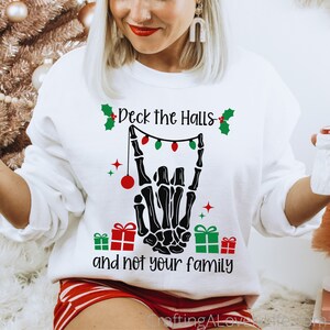 May include: White sweatshirt with a black skeleton hand making a rock and roll sign. The hand is holding a string of Christmas lights with red and green bulbs. The text "Deck the Halls - and not your family" is printed below the hand.