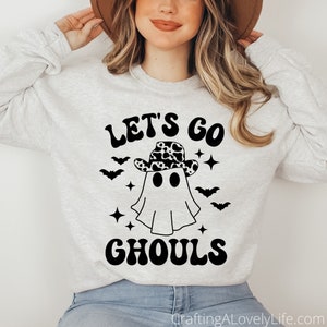 Może przedstawiać: Szara bluza z napisem "Let's Go Ghouls" w kolorze czarnym. W środku napisu znajduje się duch w kowbojskim kapeluszu z czarno-białym wzorem krowy. Duch ma dwa czarne oczy i czarne usta.