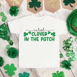 May include: White t-shirt with the text "Cutest Clover in the Patch" in black and green. The shirt is surrounded by St. Patrick's Day decorations, including shamrocks, green confetti, and a green bow tie.