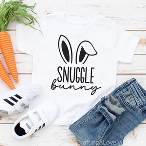 Puede incluir: Camiseta blanca con orejas de conejo negras y el texto "Snuggle Bunny" impreso en ella. La camiseta est&aacute; colocada sobre una superficie de madera blanca con un par de zapatillas blancas y unos vaqueros azules.