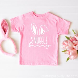 Puede incluir: Camiseta rosa con orejas de conejo blancas y el texto "SNUGGLE bunny" en ella. La camiseta est&aacute; sobre una superficie de madera blanca con una diadema de conejo rosa y flores rosas.