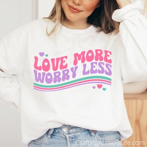 Peut inclure: Sweat-shirt blanc avec un graphisme rose, violet et turquoise qui dit "Love More Worry Less" avec des cœurs roses.