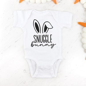 Puede incluir: Un body blanco para beb&eacute; con orejas de conejo negras y el texto "Snuggle Bunny" impreso en la parte delantera.