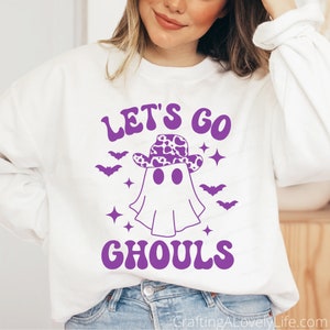 Może przedstawiać: Biała bluza z fioletowym nadrukiem graficznym przedstawiającym ducha w kowbojskim kapeluszu z wzorem krowy. Tekst "Let's go Ghouls" jest napisany fioletowym kolorem.