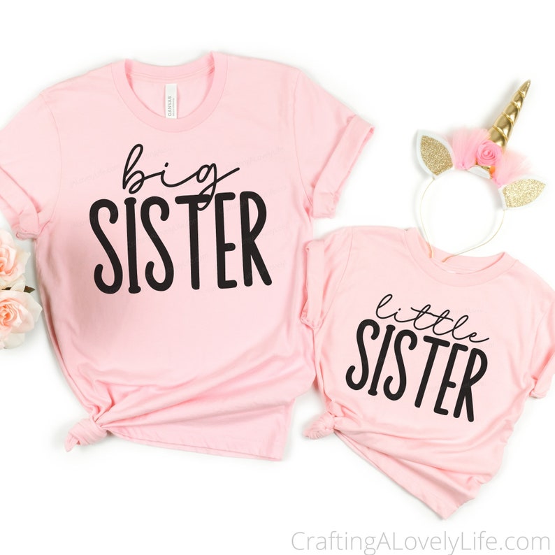 Op de afbeelding: Twee roze T-shirts met zwarte tekst. Het grotere shirt zegt "grote zus" en het kleinere shirt zegt "kleine zus".